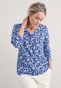 Blusenshirt mit blauem Muster und floralem Design in Weiß, aus weichem Stoff gefertigt, mit Dreiviertelärmeln und V-Ausschnitt.