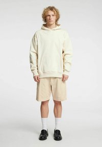 Bež kapucar z žepom spredaj, sproščen kroj, kombiniran s kremnimi kratkimi hlačami. Model nosi bele nogavice in črne čevlje brez vezalk. Enobarvna podlaga.