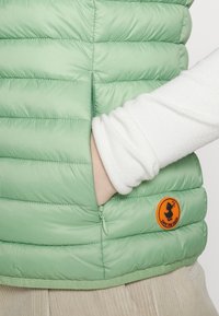 Ljust grön pufferväst med horisontell quiltad design, dragkedjeförsedd sidoficka och orange cirkulär logotyp med en anka-silhuett.