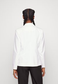 Vit blazer med skräddarsydd passform, slagkrage och en slits bak. Buren över svarta byxor, som visar en elegant design.