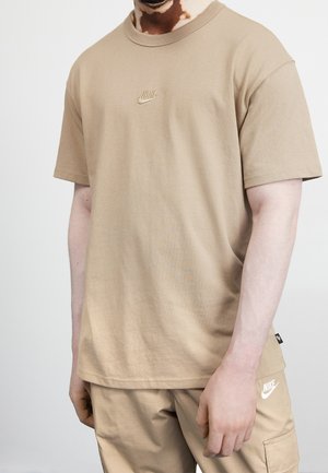 Man draagt beige Nike T-shirt en bijpassende cargobroek met zichtbare Nike-logo's, poserend tegen een effen lichte achtergrond.