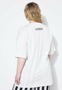 Camiseta blanca de gran tamaño hecha de algodón, con mangas cortas y un estampado gris de "LEMONS" en la parte posterior. Textura suave.