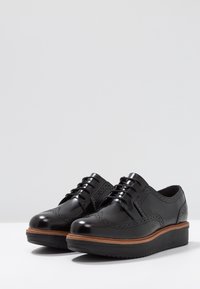 Chaussures à lacets en cuir verni noir avec des détails brogue, une semelle superposée marron et une base en caoutchouc noir légèrement surélevée.