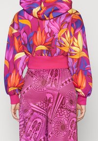 Bakifrån bild av en person med en livfull blommig hoodie med stora orange och gula blommor samt lila blad, tillsammans med rosa byxor med tropiskt mönster.