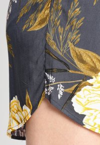 Gros plan sur un tissu foncé avec des motifs floraux jaunes et de blé, présentant un ourlet incurvé révélant la peau nue en dessous.