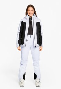 Skiausrüstung mit einer hellgrauen und schwarzen Jacke mit hohem Kragen und Reißverschlusstaschen, kombiniert mit weißen Hosen und weißen Skistiefeln.