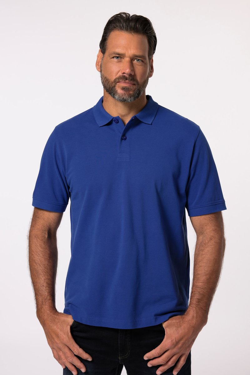 Man met baard die een blauw poloshirt met korte mouwen en zwarte broek draagt, staand met handen bij de zakken, neutrale achtergrond.