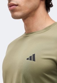 Gros plan d'une personne portant un maillot de sport texturé vert olive avec un logo noir à trois bandes sur la poitrine.