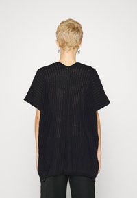 Svart stickad oversized topp med lös passform, korta ärmar, öppet mönster och texturerad yta. Bakifrån framhävs det transparenta mönstret.