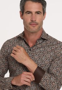 Mann mit grau meliertem Haar, der den Umschlag eines gemusterten Langarmhemds mit geometrischen Formen und braunen Innenärmeln anpasst.