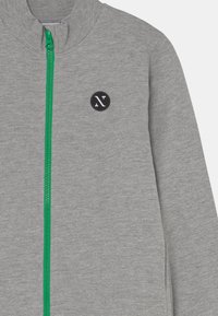 Grijze ritsjack gemaakt van zachte stof, met een groene rits, geribbelde kraag en een zwart rond logo aan de linkerkant.