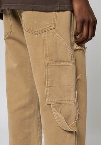 Bruna cargobyxor med flera fickor, fransade kanter och en närbild av en hand som vilar på låret. Enkel, avslappnad stil i fokus.