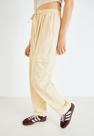 Pantalons cargo beiges avec une taille élastique et un cordon de serrage. Comprend des poches latérales et un tissu doux et léger. Associés à des baskets brunes.
