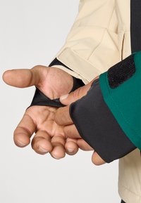 Casaco multicolorido com tecidos bege, verde e preto. Close-up de mãos a ajustar o fecho de velcro na manga, mostrando a textura e os detalhes do design.