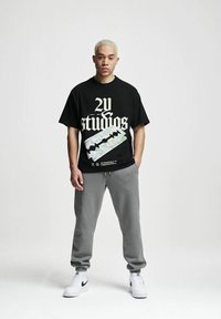 Svart bomull t-skjorte med fet hvit grafisk tekst "2Y studios" og et trykt design, sammen med grå joggebukser og hvite sko.