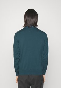 Maglione lavorato a maglia color teal con scollo rotondo e maniche lunghe, con orlo e polsini a coste, indossato sopra una camicia con colletto.