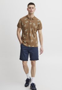 Brūns polo krekls ar zaļiem lapu rakstiem, savienots ar tumši ziliem šortiem. Modelis valkā tumši zilus apavus un baltas zeķes. Mīksta auduma, ar apkaklīti.
