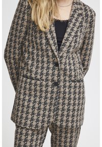 Sakó vzoru houndstooth v béžovej a čiernej farbe, s dvoma prednými vreckami, klopovými goliermi a čiernymi gombíkmi.