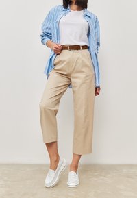 Camicia a righe azzurro chiaro, t-shirt bianca, pantaloni beige a vita alta, cintura marrone e scarpe bianche senza lacci. Texture morbide, stile informale.