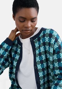 Megztinis su daugiakoloriu raštu mėlynos, žalios ir baltos spalvos, su tamsiai mėlynu dažytuvu ir penkiomis mygtukų užsegimo.