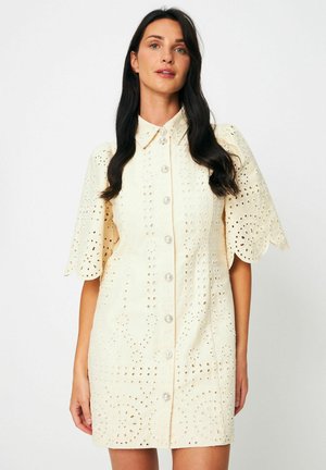 Ženska, oblečena v kremasto kratko obleko z luknjicami, valovitim robom in bisernimi gumbi spredaj, stoji ob belem ozadju.