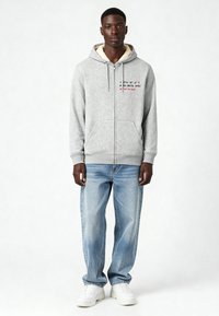 Grijze zip-up hoodie met een fleece-voering in de capuchon, voorzien van rode tekst op de borst. Gecombineerd met lichtblauwe jeans en witte sneakers.