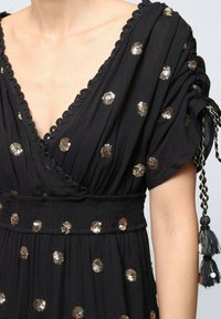 Robe noire avec des manches courtes bouffantes, un décolleté en V profond et des pois en sequins dorés. Tissu texturé avec une taille smockée et un détail en franges.