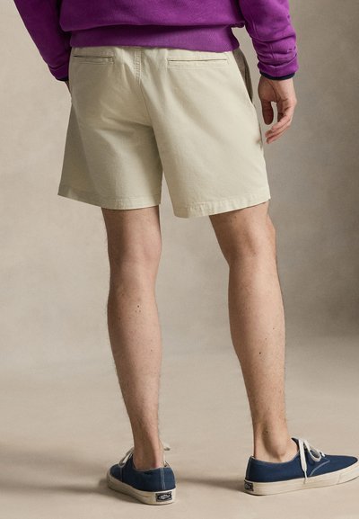 Beige cotton shorts with back pockets, featuring a straight cut and hem. Model pairs them with blue canvas shoes. Focus on legs visible.