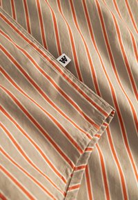 Tissu beige avec des rayures verticales orange et blanches. Étiquette notable cousue dans le coin. Texture lisse, bords soigneusement finis.