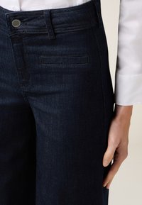 Jeans de mezclilla oscura con un diseño ajustado, textura suave, costuras sutiles y un cierre de botón clásico con un detalle de bolsillo lateral.