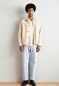 Beige jacket over a textured cream shirt, light blue jeans, and black ankle boots, against a plain background. Focus on fit and color contrast.