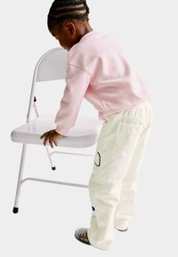 Niño vestido con una sudadera rosa y pantalones blancos con diseños bordados, apoyado en una silla plegable blanca, con calzado texturizado.