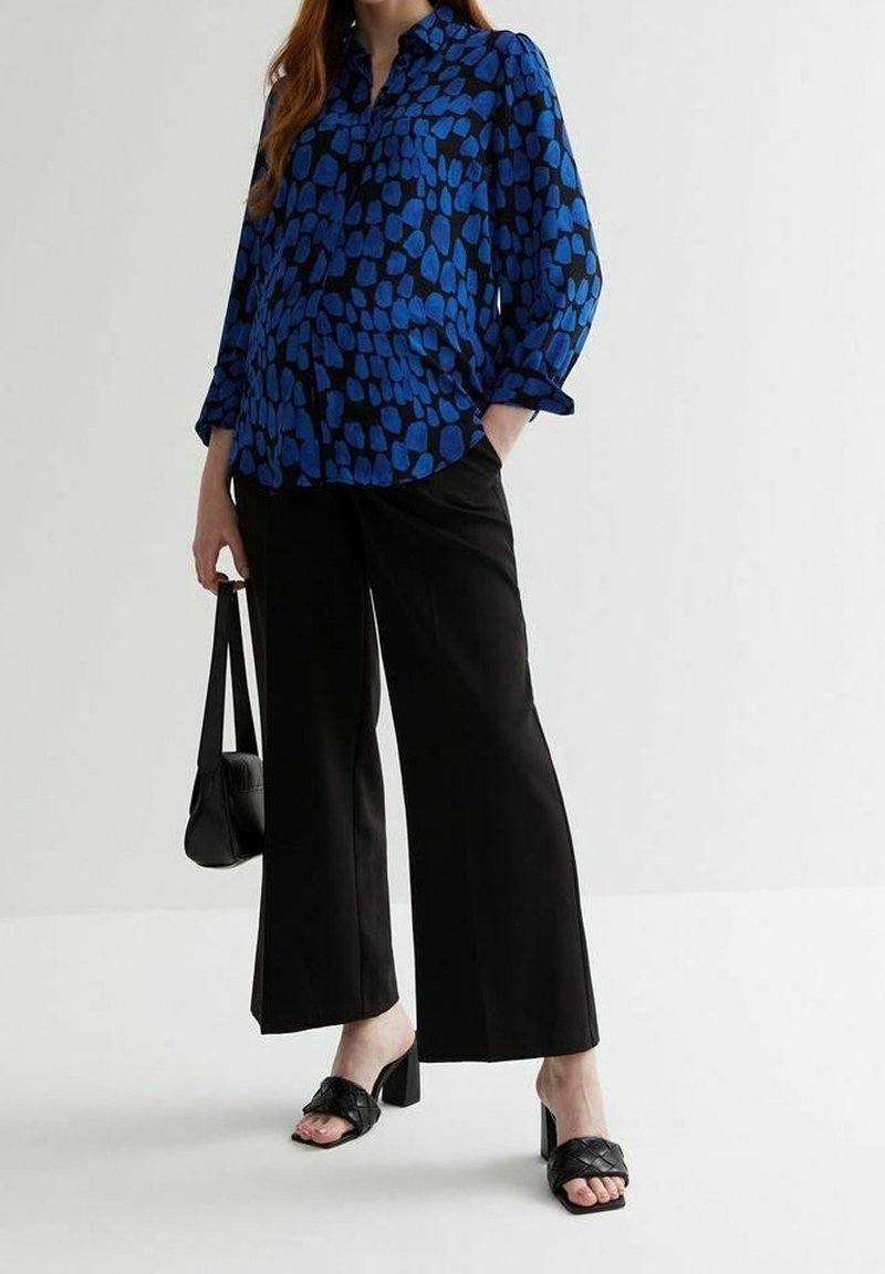 Femme portant un chemisier bleu à motifs, un pantalon noir à jambes larges, des sandales noires à talons et tenant un petit sac à main noir.