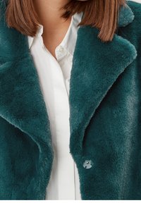 Zielona sztuczna futrzana kurtka z kołnierzem w kształcie litery V i pojedynczym zapięciem na guzik. Miękka tekstura, luźny krój, noszona na białej koszuli.