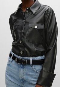 Chemise noire en cuir avec un col classique, deux poches poitrine, des boutons-pression argentés et des manches longues, associée à un jean et une ceinture noire.