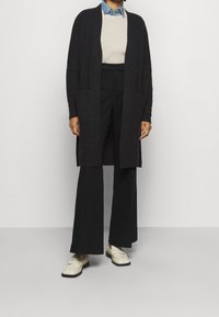 Dlouhý otevřený kardigan v tmavě šedém pleteném materiálu, se dvěma předními kapsami, přetažený přes béžový svetr a džínovou košili, doplněný černými kalhotami a mokasíny.