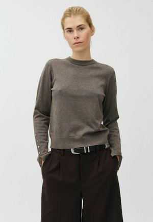 Femme aux cheveux blonds attachés derrière la tête portant un pull gris à manches longues avec des boutons sur les manches et un pantalon marron foncé avec une ceinture noire.