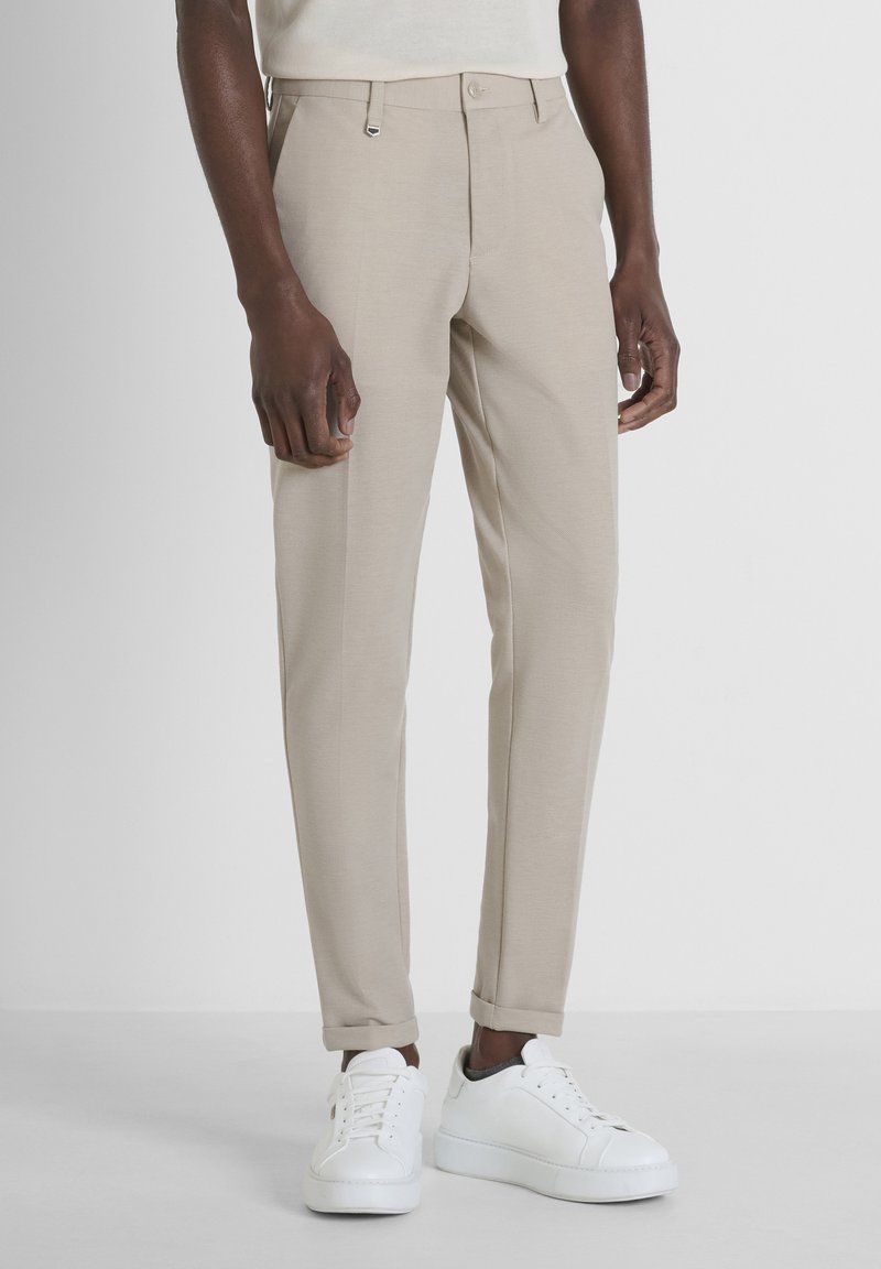 Pantaloni beige su misura con vestibilità slim, realizzati in tessuto liscio. Presentano una chiusura a bottoni, passanti per la cintura e orli arrotolati. Indossati con scarpe da ginnastica bianche.