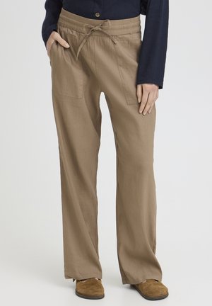 Személy bézs színű, széles szárú nadrágot visel, húzózsinóros derékrésszel és oldalzsebekkel, tengerészkék inget, valamint barna szarvasbőr cipőt, egyik kezét a zsebében tartva áll.