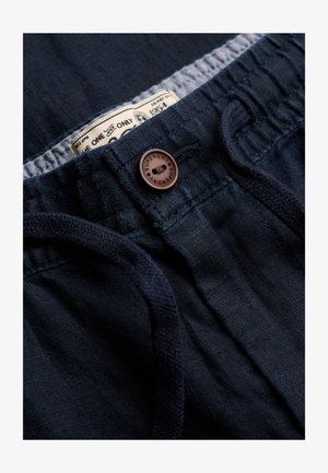 Zbliżenie na ciemnogranatowe lniane spodnie z elastycznym pasem, sznurkiem, brązowym guzikiem z napisem „Superdry” oraz częściowo widoczną białą metką z tkaniny w środku.