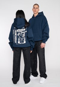 Haut à capuche zippé bleu marine avec un imprimé graphique au dos, associé à un jean large noir et des baskets blanches. Texture lisse dans l'ensemble.