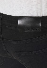 Černé džíny z denimu s koženou nášivkou označenou "True Religion", s detailním záběrem pasu a zadní kapsy.