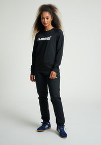Felpa nera con logo "hummel" bianco, abbinata a pantaloni jogger neri. Il modello indossa scarpe sportive blu con suola in gomma. Capelli acconciati in ricci.