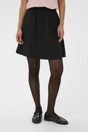 Nő fekete redős szoknyában, fekete áttetsző harisnyában és fekete lakkbőr balerinacipőben, kezei lazán az oldalán pihennek.