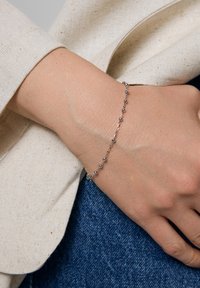 Srebrna bransoletka z małymi okrągłymi koralikami, cechująca się delikatnym wzorem łańcuszka. Noszona na dłoni na tle jasnej tkaniny i dżinsu.