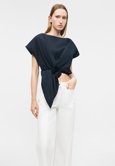 Γυναίκα που φορά μαύρο κοντομάνικο τοπ δεμένο στη μέση και λευκό παντελόνι ψηλόμεσο με φαρδιά μπατζάκια, στέκεται με το ένα χέρι στην τσέπη.