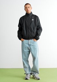Sort vindjakke med høj krave og elastiske manchetter, parret med lyseblå baggy jeans og grå sneakers. Enkel, funktionel design.