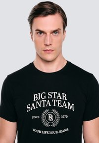 Černé bavlněné tričko s bílým grafickým textem: "BIG STAR SANTA TEAM" a logem, kulatý výstřih, krátké rukávy, hladká textura.