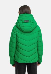 Personne portant une veste matelassée verte avec capuche, vue de dos, se tenant devant un fond clair et uni.
