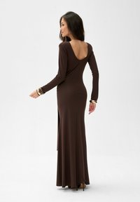 Brązowa sukienka z długim rękawem, z głębokim dekoltem na plecach, dopasowana sylwetka, rozcięcie po boku i długość do ziemi, uzupełniona wysokimi obcasami i złotymi bransoletkami.